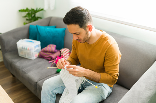 Adult man looking happy while knitting a wool scarf. Man in his 30s sitting on the sofa and practicing his favorite hobby