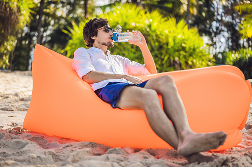 Summer lifestyle portrait of men sitting on the orange inflatable sofa drinking water on the beach of tropical island. Relaxing and enjoying life on air bed.