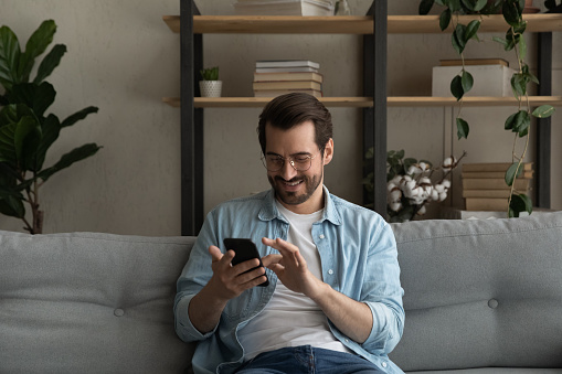 Content young man relax on sofa use smartphone spend free time online in social network chat at web app play easy game online. Smiling guy hold modern cell dial number on screen make call to friend