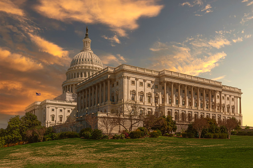 South Wing (the House of Representatives wing) of the US Capitol Building, Washington DC, USA. Green bushes, grass and trees are in foreground. Orange, Yellow and Blue Sunset Sky with Puffy Clouds is in background.