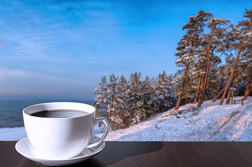 Cup of coffee on the table with view of snowy beach, Baltic sea and covered in snow fir and pine trees on the hill.