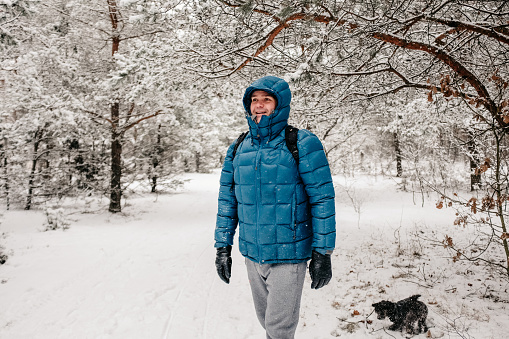 January 30, 2021 - Poland: Relaxed, cheerful, attractive, handsome mature man standing in a winter forest covered with deep snow wearing a blue jacket, hood, looking away laughing, mouth open, arms at sides, dog running.
