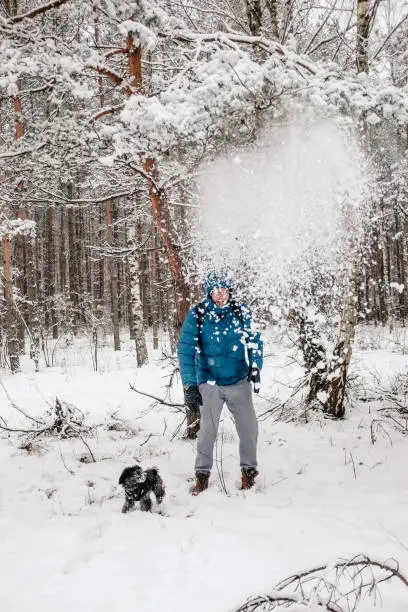 January 30, 2021 - Poland: Man in blue jacket standing under a snow shower after shaking the pine tree in fairy tale idyllic winter forest with a puppy dog.