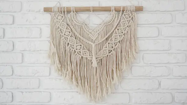 Wall panel in the style of Boho made of cotton threads in natural color on a white brick wall. Beautiful boho macrame wall panel for a cozy atmosphere