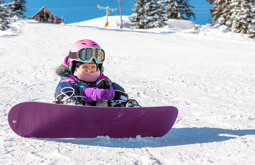 Little girl snowboarding at ski resort in sunny winter day. Portrait of kid in sportswear with equipment resting on ski slope
