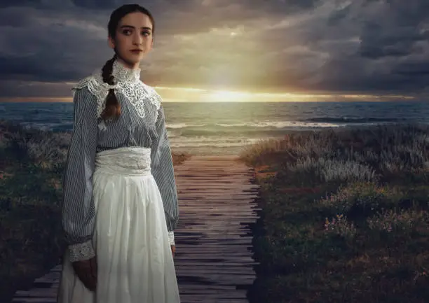 Young victorian girl in white dress and blue striped blouse on the coast at sunset.