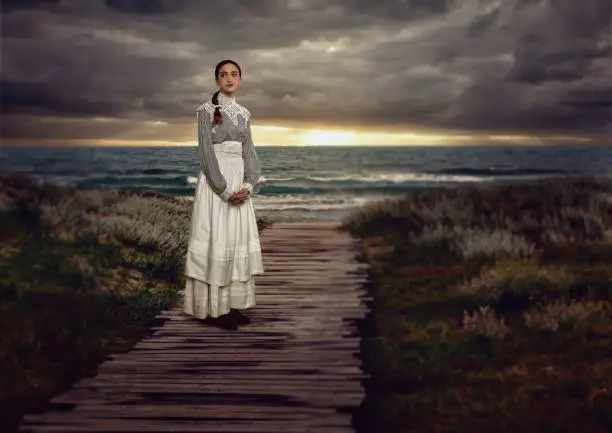 Young victorian girl in white dress and blue striped blouse on a boardwalk at the coast at sunset.