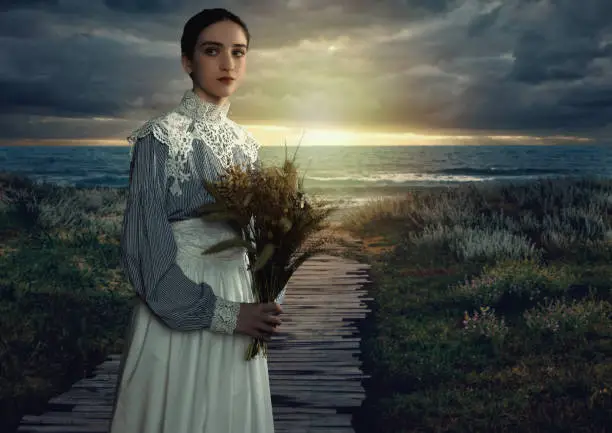 Young victorian girl holding dried flowers in white dress and blue striped blouse at the coast at sunset.