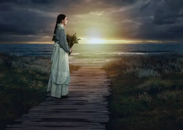 Young victorian girl holding dried flowers in white dress and blue striped blouse on a boardwalk at the coast at sunset.