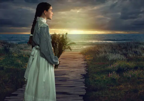 Young victorian girl holding dried flowers in white dress and blue striped blouse at the coast at sunset.