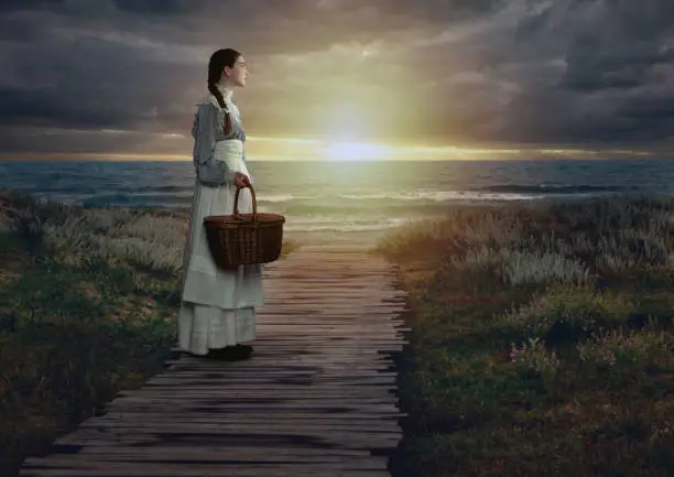 Victorian girl with a wicker basket in white dress and blue striped blouse on a seaside promenade at sunset.