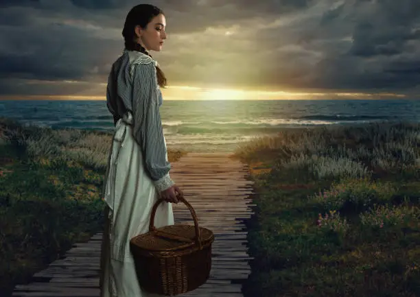 Victorian girl with a wicker basket in white dress and blue striped blouse at seaside at sunset.