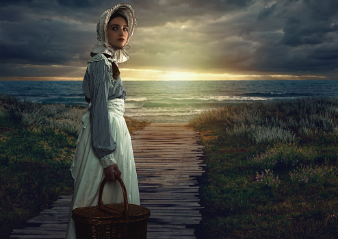 Victorian girl with hood and wicker basket in white dress and blue striped blouse on the coast at sunset.