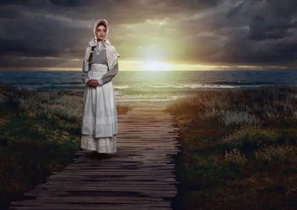 Victorian girl with a cap on in a white dress and blue striped blouse on the coast at sunset.