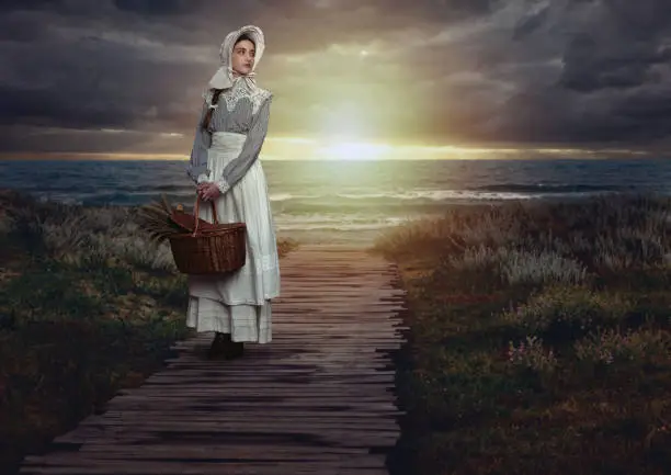 Victorian girl with a hood and a wicker basket containing dried flowers in a white dress and blue striped blouse on the coast at sunset.