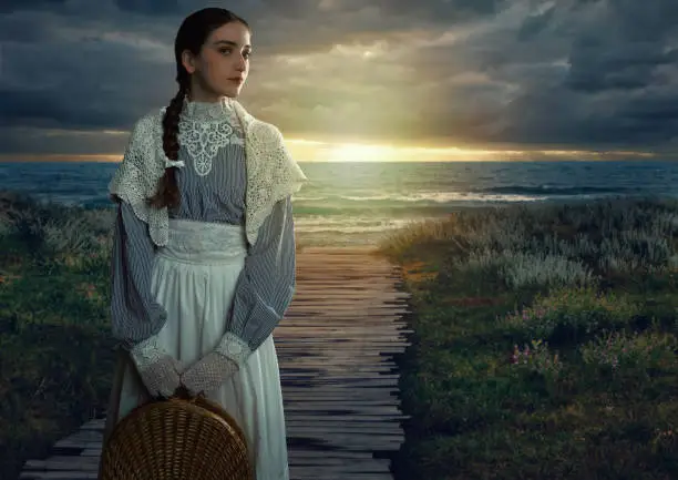 Victorian girl with a hood and a wicker basket in her hands dressed in a white dress and blue striped blouse stands on the seashore at sunset.