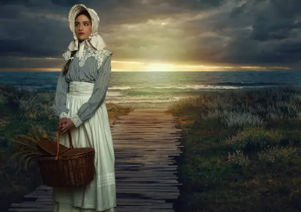 Victorian girl with a hood and a wicker basket containing dried flowers in a white dress and blue striped blouse on the coast at sunset.