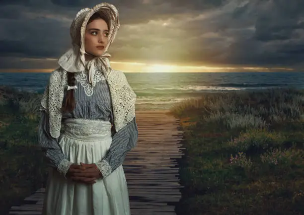 Victorian girl with a cap on in a white dress and blue striped blouse on the coast at sunset.