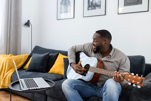 Young African American male musician or teacher playing guitar during an online concert lesson at home while isolated, sitting on the sofa, using laptop to stream record music, chatting with friends