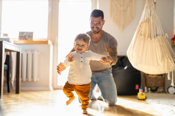 feliz padre ayudando a su hijo pequeño caminando en la sala de estar - hijos fotografías e imágenes de stock