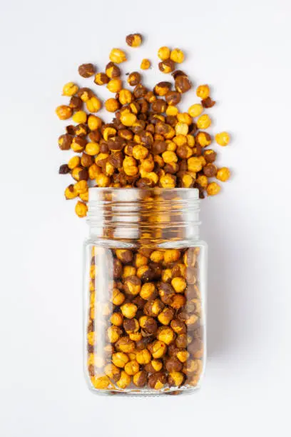 Crunchy Roasted Chana Masala spilled out and in a glass jar, made with Bengal Grams or Chickpeas. Indian spicy snacks (Namkeen),  Top view, over the white background.