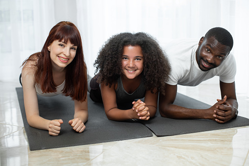 At plank. Handsome multiracial parents doing plank with their daughter at the yoga mat. Parents including child in activity. Family quarantine, domestic life in self-isolation concept