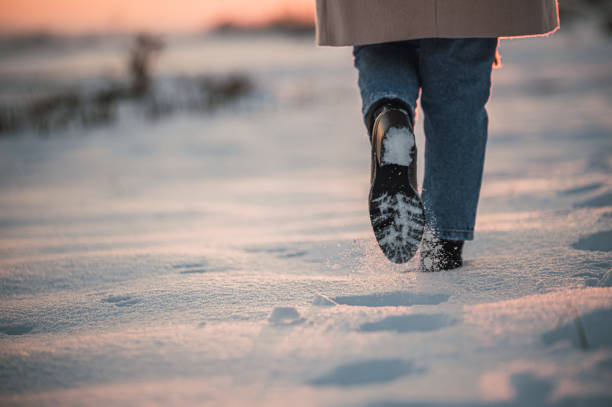 Back view of crop unrecognizable woman in warm clothes and boots leaving footprints while walking alone on fresh white snow in winter countryside at sunset time