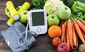 Blood pressure monitor, dumbbells and fresh fruits with vegetables against wooden table.