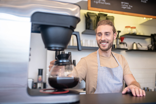 Coffee, cafe. Smiling young adult man in apron with coffee pot near coffee maker behind counter in cafe