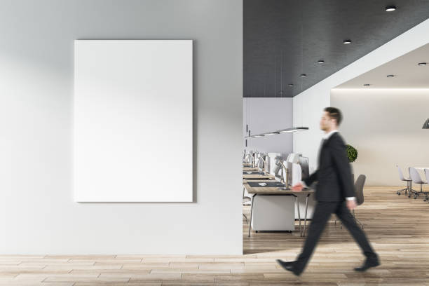 istock Businessman goes to blank white poster on light grey wall in modern open space office with light furniture and wooden floor 1304086601