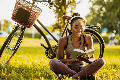 Photo of a young African American woman is wearing wireless headphones, she is sitting on the grass next to her bicycle