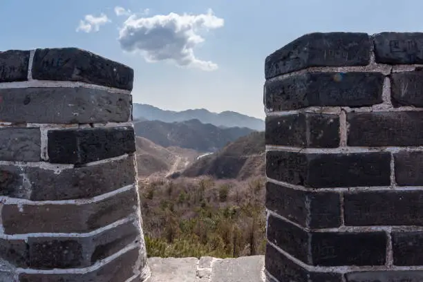 Beijing, China - April 28, 2010: Great Wall of China. Look through dark stone battlements upon will hilly landscape with stretch of wall in distance under blue cloudscape.
