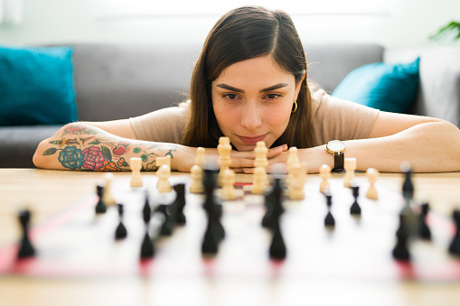 Attractive woman looking at the chess pieces on the board and thinking about a move to win the game. Thoughtful woman enjoying a relaxing day at home