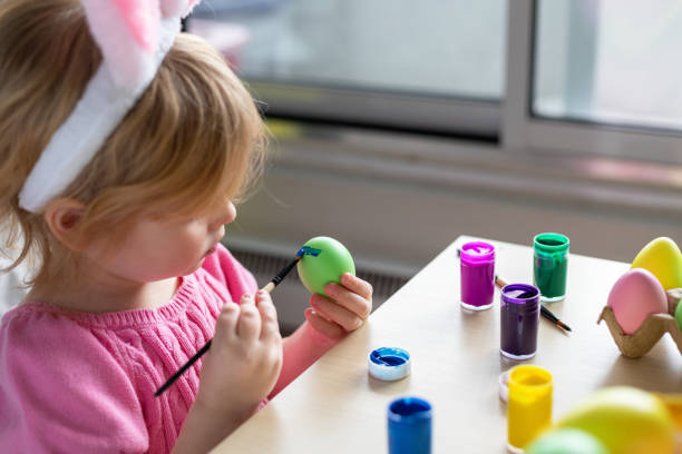 mestiere per le vacanze di pasqua a casa. bambina nelle orecchie da coniglietto pasquale che dipinge uova colorate. - easter easter egg paintbrush holding foto e immagini stock