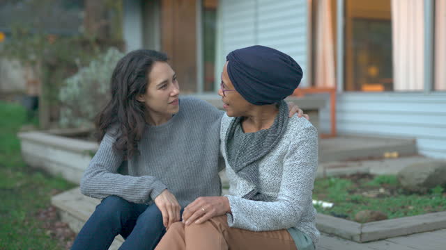 A senior woman with cancer is embraced and comforted by her adult daughter as they sit outside on a fall evening. The mother is smiling and laughing while the daughter is squeezing her mother affectionately and smiling as well.