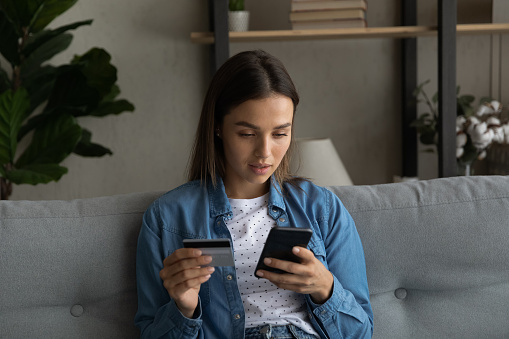 Concentrated millennial female sit on sofa at living room hold plastic credit card provide cashless payment transaction via electronic bank. Focused young lady check card account balance at phone app