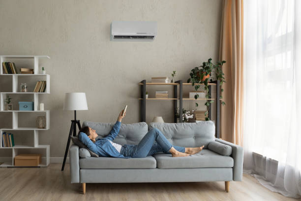 Happy young lady regulate climate at home using ac device Glad millennial female buyer of air conditioner relax on sofa hold controller breath cool fresh air despite of hot summer day outside. Happy young lady regulate climate at home using modern ac device cool climate stock pictures, royalty-free photos & images