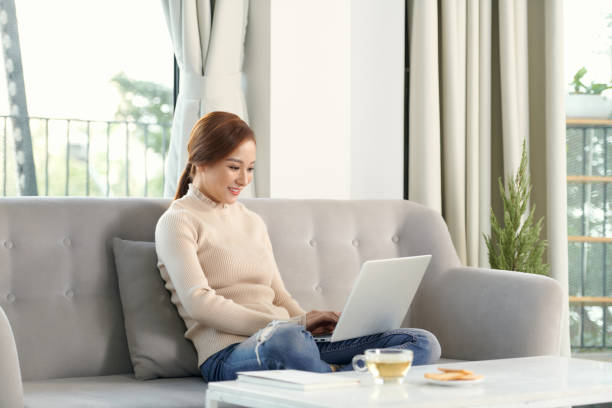 istock Smiling young woman using laptop, sitting on couch at home, 1304133943