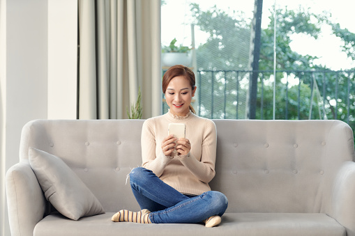 Happy young Vietnamese woman relax in living room browse wireless internet on cellphone gadget.