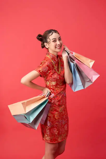 beautiful Asian woman wearing traditional cheongsam qipao dress showing shopping bags and gold necklace on red background for Chinese new year shopping concepts.