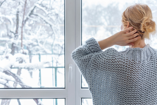 A woman dressed in a cozy sweater looks out the window straightening her disheveled hair with her hand