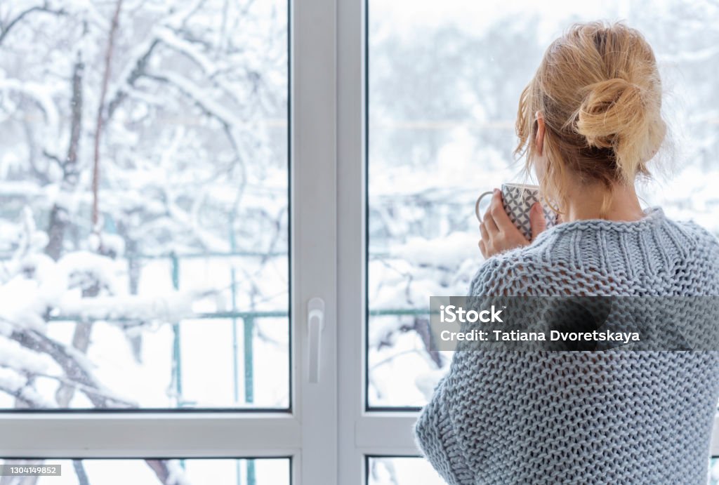 A woman looks out of the window at the snow-covered outdoors A woman in a cozy sweater stands at the picture window with a cup of drink in her hands Winter Stock Photo