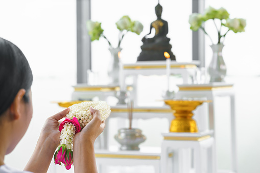 flowers garland in hands of religious asian buddhist woman worship and pray with faith to buddha statue at home