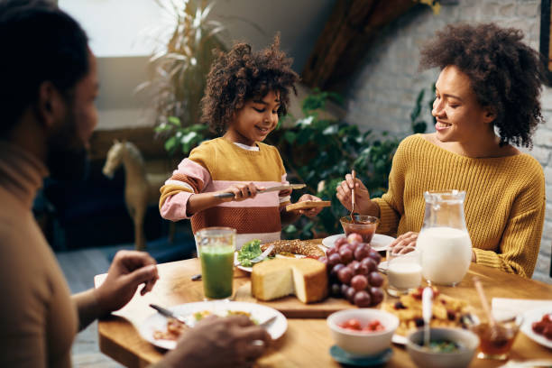 fille afro-américaine heureuse ayant le petit déjeuner avec ses parents à la table à manger. - butter bread breakfast table photos et images de collection