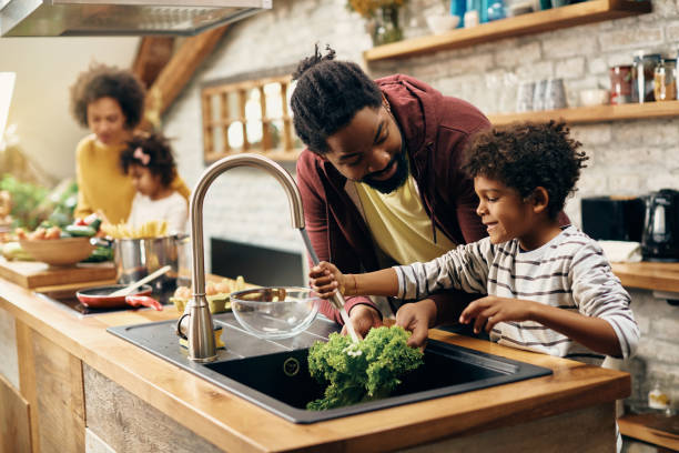 Happy black father and son cleaning vegetables while preparing food in the kitchen. Happy African American father and son washing salad at kitchen sink while making a meal together. Mother and daughter are in the background. kitchen sink stock pictures, royalty-free photos & images