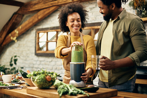 Happy black couple blending fresh vegetables and making smoothie in the kitchen.