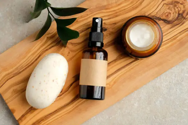 Photo of Set of natural organic SPA beauty products on wooden board. Homemade soap, moisturizer cream jar, amber glass spray bottle, green leaf on wooden board.