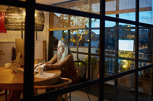 Partial view of gray-haired Caucasian woman wearing eyeglasses and casual clothing photographed through sun room window at end of workday.
