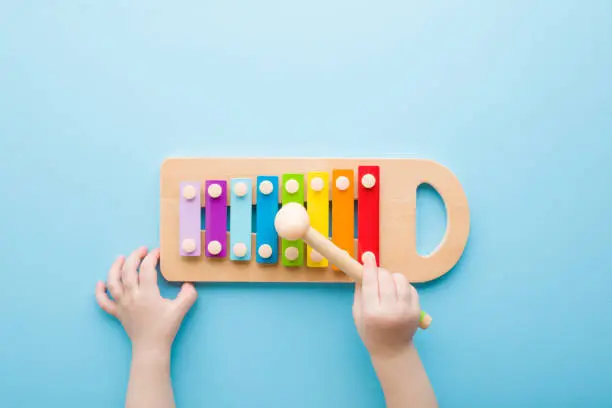 Photo of Baby hand holding hammer and playing colorful xylophone on light blue table background. Closeup. Music toy instrument of development for little kids. Point of view shot. Top down view.