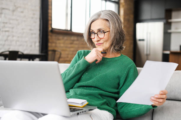 Serious senior accountant sitting on the living room and looking at the laptop stock photo